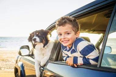 Child and dog smiling out of a car window at the beach.