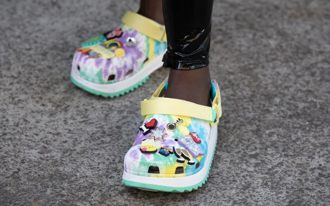 Colorful clogs with pins and designs, worn with black leggings, on a concrete surface.