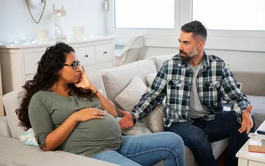 Pregnant woman and man sitting on a couch, engaged in a serious conversation inside a brightly lit living room.