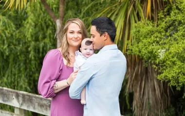 Family posing outdoors with mother in purple dress, father in blue shirt, holding a smiling baby.