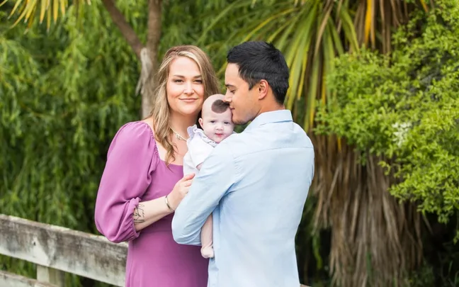Family posing outdoors with mother in purple dress, father in blue shirt, holding a smiling baby.
