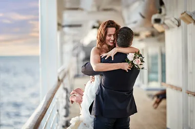Couple embracing joyfully on a ship's deck, holding a bouquet, with the ocean in the background.