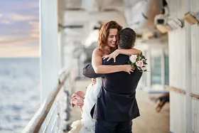 Couple embracing joyfully on a ship's deck, holding a bouquet, with the ocean in the background.