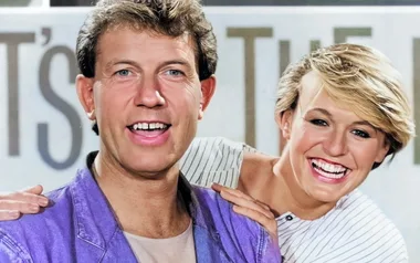 Two people smiling, one wearing a purple jacket, in front of a blurred background with partial text.