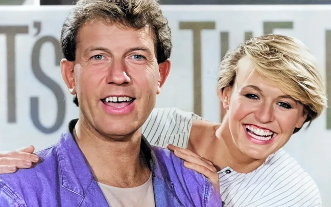 Two people smiling, one wearing a purple jacket, in front of a blurred background with partial text.