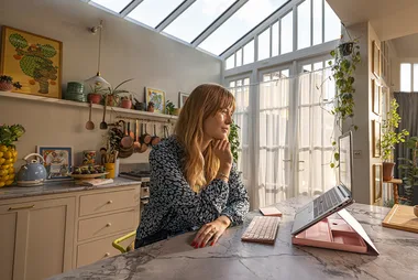Woman in patterned top works on a laptop at a marble kitchen counter with plants and artwork in the background.