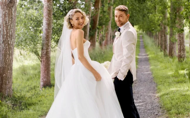 A bride in a white gown and a groom in a light suit smile at each other on a tree-lined path.