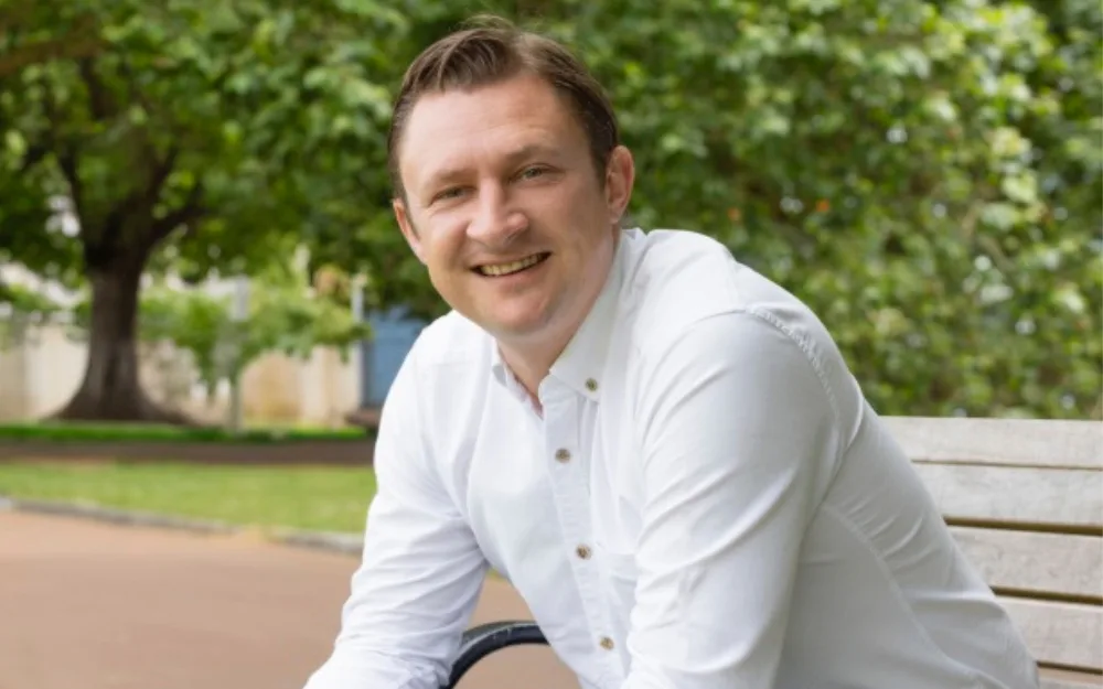Man in a white shirt sitting on a bench in a park setting with trees in the background.