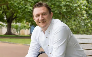 Man in a white shirt sitting on a bench in a park setting with trees in the background.