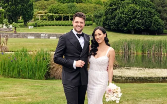 A couple in wedding attire poses outdoors near a pond with lush greenery in the background.
