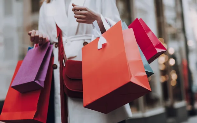 Person holding multiple colorful shopping bags outside stores.