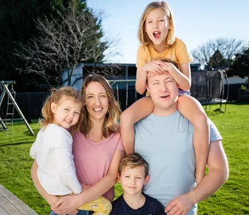 A family of five smiling in their backyard; a woman holds a child, and a man carries another on his shoulders.
