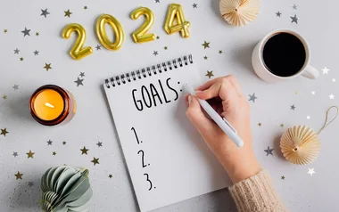 Planning 2024 goals, hand writes in notebook with candle, coffee, decorations, and gold numbers.