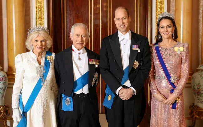 A royal group in elegant attire with sashes and tiaras, standing in a formal setting.