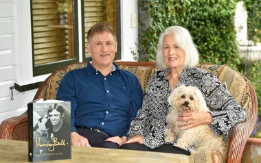 A man and woman sit on a porch with a small dog. A book titled "Mason Ball" is on the table.