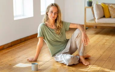 Person sitting on wooden floor in casual clothes with a mug nearby, soft lighting from windows.