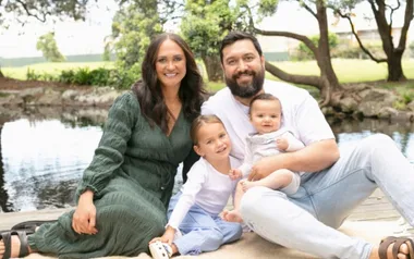 A family of four sitting by a pond, with trees in the background, smiling and wearing casual clothes.