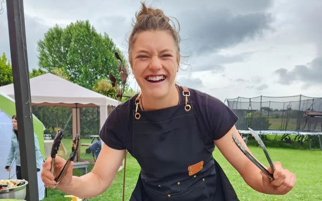 Person smiling outdoors holding barbecue tongs, wearing a black apron, with a garden and tents in the background.