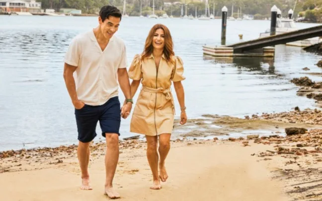 A couple walking barefoot on a beach near the water, smiling and holding hands.
