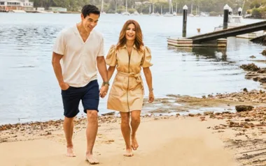 A couple walking barefoot on a beach near the water, smiling and holding hands.