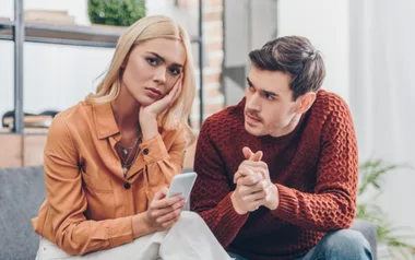 Man in red sweater talks to woman in orange shirt holding a phone, both seated on a couch in a cozy room.