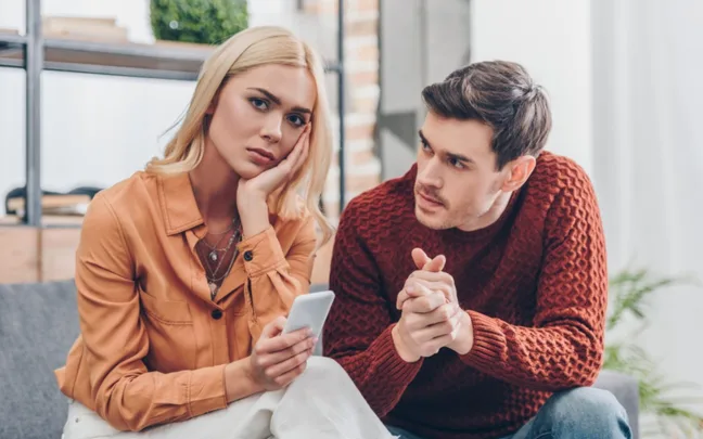 Man in red sweater talks to woman in orange shirt holding a phone, both seated on a couch in a cozy room.