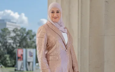 Woman in a light hijab and sequin jacket smiles while standing outdoors near a column.