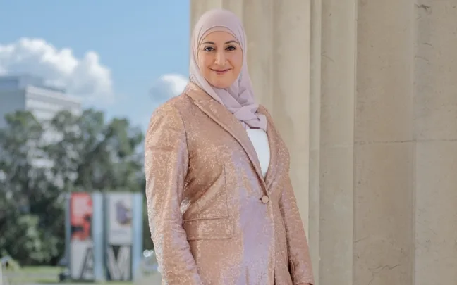 Woman in a light hijab and sequin jacket smiles while standing outdoors near a column.