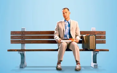 A man in a light suit sits on a bench with a suitcase, holding a box of chocolates against a blue background.