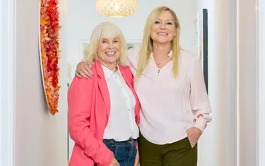 Two smiling women standing in a brightly lit hallway, one wearing a pink blazer and the other a pale pink blouse.
