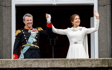 A man and woman in formal attire stand on a balcony holding hands, smiling and waving to a crowd.
