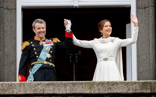 A man and woman in formal attire stand on a balcony holding hands, smiling and waving to a crowd.