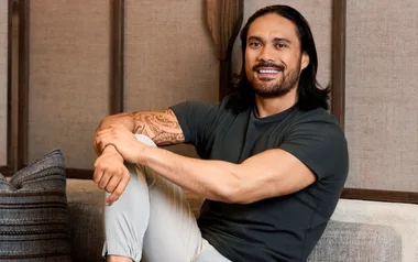 A person with long dark hair, sitting on a couch, smiling, wearing a dark shirt and light pants.