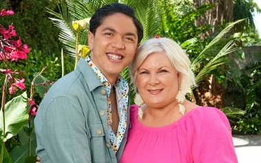 Two people smiling and posing together in a lush green garden, with one wearing a blue jacket and the other in a pink top.