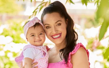 A woman and a baby smiling happily outdoors, surrounded by greenery and sunlight.