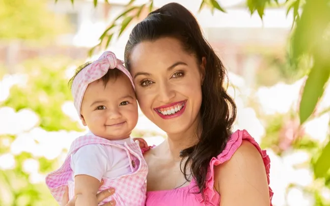 A woman and a baby smiling happily outdoors, surrounded by greenery and sunlight.