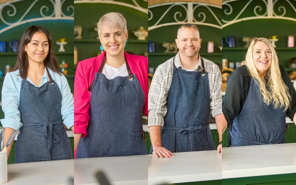 Four contestants in aprons smile at a kitchen counter in the set of a baking competition show.