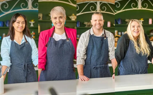 Four contestants in aprons smile at a kitchen counter in the set of a baking competition show.