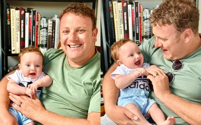 Man holding a smiling baby, both wearing casual clothes, with bookshelves in the background.