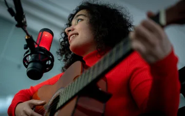 Musician in a red sweater plays guitar and sings into a glowing red microphone.