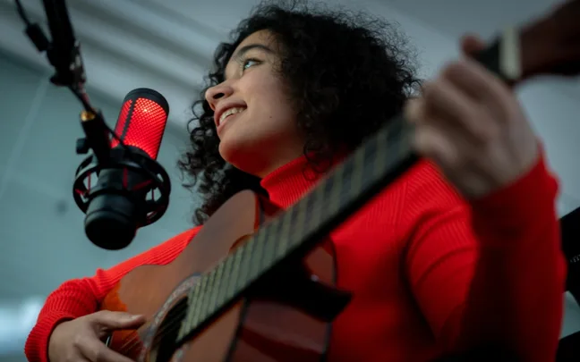 Musician in a red sweater plays guitar and sings into a glowing red microphone.