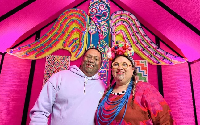 Two people smiling in front of colorful, pink-themed textile art background with intricate patterns.