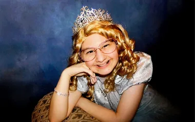 A young person in a fancy dress, curly blonde wig, crown, and glasses, smiling against a dark background.
