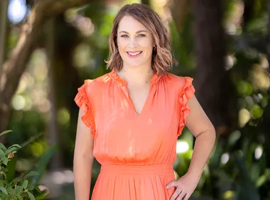 Woman in an orange dress smiling outdoors with greenery in the background.