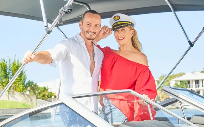 Man in white shirt and woman in red top with sailor hat, smiling on a boat.