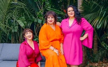 Three women smiling in colorful dresses, surrounded by lush green foliage.