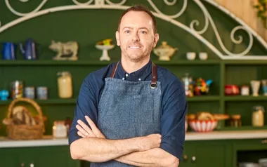 Man in denim apron standing with arms crossed, in a rustic kitchen setting with green shelves and decorative items.