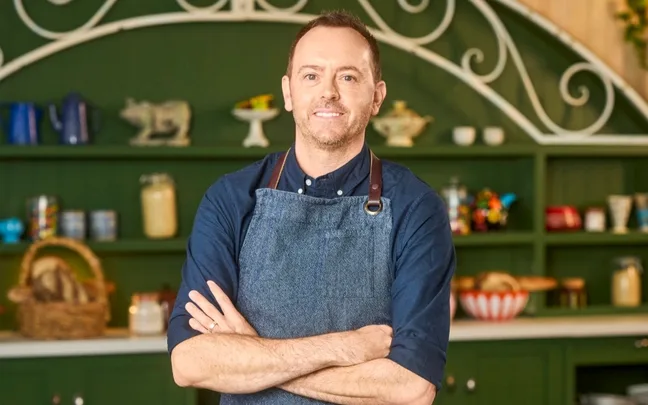 Man in denim apron standing with arms crossed, in a rustic kitchen setting with green shelves and decorative items.