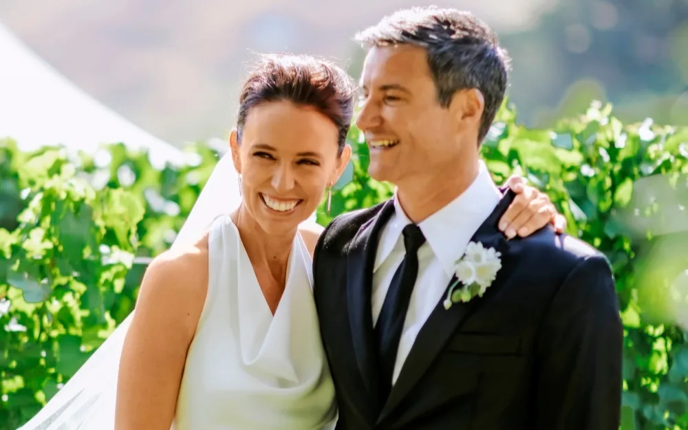 A bride and groom smiling outdoors, surrounded by greenery.