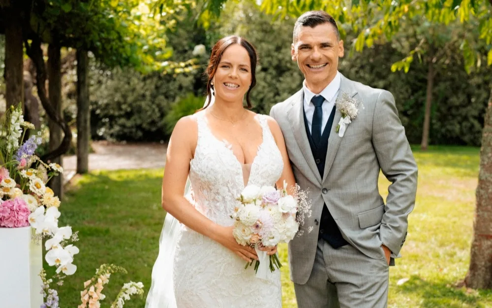 A bride in a white gown and a groom in a gray suit smiling outdoors in a garden setting.